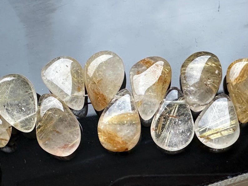May include: A strand of teardrop-shaped beads made of clear quartz with gold inclusions. The beads are strung together with a silver wire.