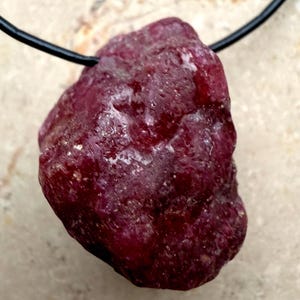 Könnte beinhalten: Ein Anhänger aus rohem Rubin. Der tiefrote Stein hängt an einem schwarzen Band. Der Rubin hat eine raue, strukturierte Oberfläche mit helleren Einschlüssen, die seine natürliche Form zeigen. Ein einzigartiges Schmuckstück.