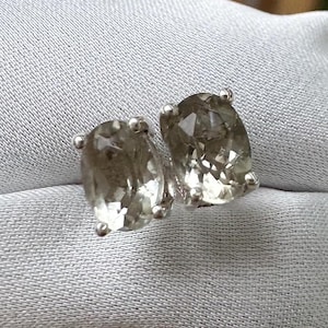 May include: A pair of oval-cut, light green gemstone stud earrings. Each earring features a faceted, translucent stone set in a silver-colored metal setting. The earrings are displayed on a light gray fabric.