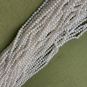 Könnte beinhalten: Stränge aus kleinen, weißen Kunstperlen liegen auf einem grünen Untergrund. Die Perlen sind gleichmäßig groß und erzeugen einen zarten, eleganten Eindruck. Ideal für Schmuckherstellung oder Bastelprojekte.