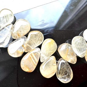 May include: A strand of golden rutilated quartz gemstone beads. The beads are teardrop shaped and have a translucent, light brown color with gold inclusions.