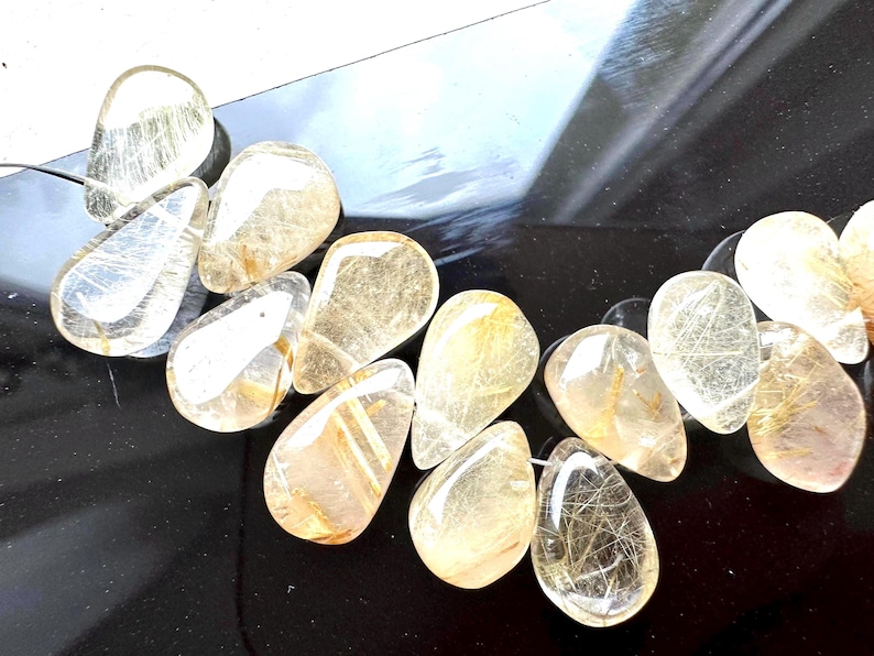 May include: A strand of golden rutilated quartz gemstone beads. The beads are teardrop shaped and have a translucent, light brown color with gold inclusions.
