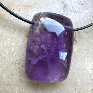 Pode incluir: Um pingente de ametista retangular polido, com um tom roxo profundo. A pedra preciosa é presa a um cordão preto, criando um colar simples e elegante. A superfície do pingente reflete a luz, mostrando sua textura natural e variações de cor.