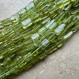 Könnte beinhalten: Nahaufnahme eines Strangs transluzenter, hellgrüner Peridot-Perlen. Jede Perle ist rechteckig mit abgerundeten Kanten und aufgefädelt. Die Perlen haben ein glasartiges Aussehen und reflektieren das Licht. Der Hintergrund ist eine neutrale, strukturierte Oberfläche.