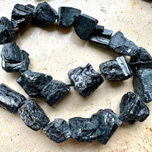 Può includere: Una collana di perle di turmalina nera. Le perle sono grezze e di forma irregolare, di un colore scuro, quasi nero. Le perle sono infilate su un cordoncino nero.