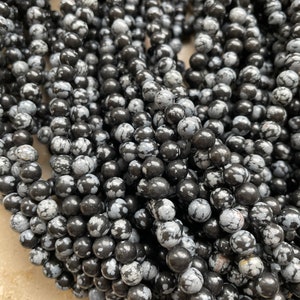 Peut inclure: Une collection de perles rondes en obsidienne flocon de neige présentant une surface noire polie avec des motifs tachetés gris et blancs distincts. Ces perles sphériques en pierre sont disposées en plusieurs rangs, révélant une texture lisse.