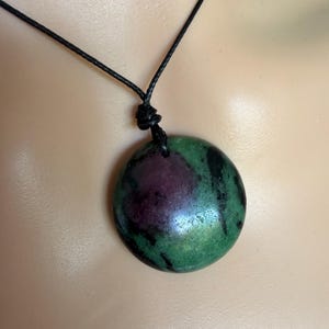 May include: A round, polished pendant featuring a mottled pattern of deep green and dark black mineral inclusions with a prominent circular patch of magenta-colored stone. The pendant is suspended from a simple, thin black cord with a knotted detail.