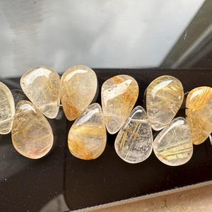 May include: A strand of teardrop-shaped, clear quartz beads with gold inclusions. The beads are faceted and have a natural, raw look.