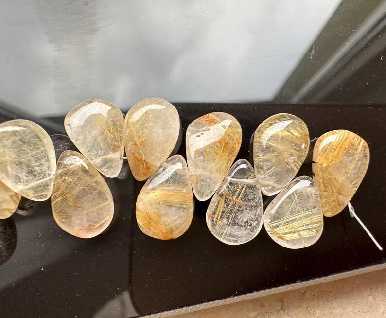 May include: A strand of teardrop-shaped, clear quartz beads with gold inclusions. The beads are faceted and have a natural, raw look.