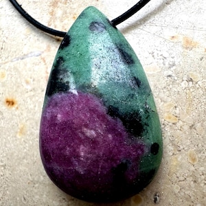 Può includere: Una collana con pendente a goccia con una pietra lucida che mostra tonalità verdi, nere e viola intenso. La pietra è sospesa a un cordino nero, creando un contrasto sorprendente. Il motivo cromatico unico del pendente ne aumenta l'appeal.