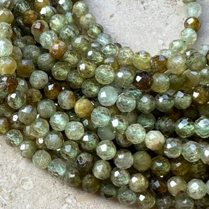 Pode incluir: Um fio de contas de pedra preciosa verde facetadas. As contas são redondas e têm uma forma ligeiramente irregular. São de cor verde claro com um toque de castanho.