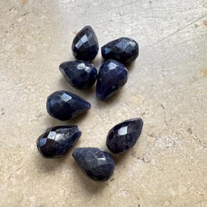 Può includere: Una collezione di sette gemme sfaccettate a forma di goccia, di un profondo colore indaco. Le gemme sono disposte su una superficie beige chiaro, ognuna cattura la luce con molteplici sfaccettature. Ideale per la creazione di gioielli.