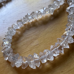 Peut inclure: Collier de perles en cristal de quartz clair, chacune de forme irrégulière et facettée. Les perles sont enfilées sur un cordon blanc. Le collier est présenté sur une surface en bois. Les perles sont translucides.
