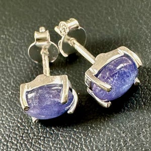 Könnte beinhalten: Ein Paar silberne Ohrstecker mit jeweils einem violetten Edelstein. Die Edelsteine sind oval geformt und haben eine glitzernde Oberfläche.