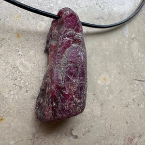 Könnte beinhalten: Ein Anhänger aus rohem, tiefrotem Rubin, an einer schwarzen Kordel befestigt. Der Rubin hat eine raue, natürliche Textur mit silbernen Akzenten und eine leicht längliche Form. Die Kordel scheint aus Leder oder einem ähnlichen Material zu sein.