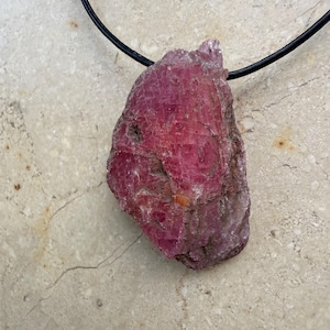 May include: A raw, deep pink ruby pendant suspended from a thin black cord. The gemstone features a jagged, unpolished surface with natural crystalline textures and variations in color, ranging from dark magenta to lighter translucent pink hues.