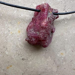 Può includere: Un ciondolo con pietra preziosa di rubino grezzo e non tagliato. Il rubino è di un colore rosso intenso e ricco, con una superficie ruvida e strutturata. È infilato su un cordino nero, creando una collana semplice e naturale. La gemma è il punto focale.