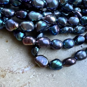 Könnte beinhalten: Nahaufnahme von mehreren Strängen irisierender, dunkel gefärbter Barockperlen. Die Perlen zeigen eine Reihe von Farben, darunter tiefes Blau, Lila und Grün, mit einer glänzenden, unregelmäßigen Oberfläche. Die Perlen sind aufgefädelt, was auf Schmuck hindeutet.