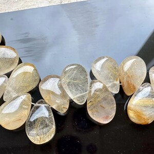 May include: A strand of teardrop-shaped, clear quartz beads with gold inclusions. The beads are arranged in a curved line on a black surface.