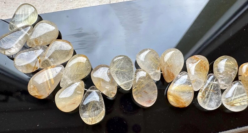 May include: A strand of teardrop-shaped, clear quartz beads with gold inclusions. The beads are arranged in a curved line on a black surface.