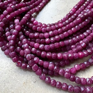 Może przedstawiać: Szereg głębokich różowych fasetowanych koralików z kamieni szlachetnych. Koraliki są małe i okrągłe, o lekko nieregularnym kształcie. Koraliki są nawleczone na przezroczystą nić.