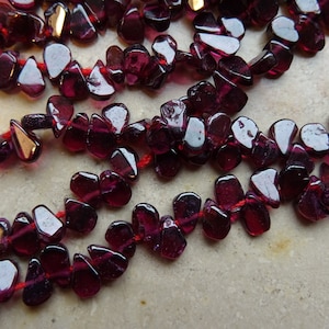 Könnte beinhalten: Eine Strähne tief roter Edelsteinperlen, wahrscheinlich Granat, auf rotem Faden aufgereiht. Die Perlen sind unregelmäßig geformt und haben eine glänzende Oberfläche.