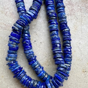 Op de afbeelding: Een streng diepblauwe lapis lazuli kralen, elke kraal is een platte, ronde schijf met een licht onregelmatige vorm. De kralen zijn aan elkaar geregen om een ​​lange ketting te vormen.