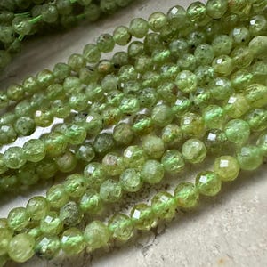 Op de afbeelding: Close-up van strengen lichtgroene edelsteen kralen. De kralen zijn facetgeslepen, rond en hebben een getextureerd oppervlak. Ze zijn geregen op een groene draad. Elke kraal is ongeveer 6 mm in diameter.