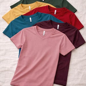 May include: A stack of six short-sleeved v-neck t-shirts in various solid colors including dark green, yellow, red, teal, maroon, and dusty pink. Each shirt features a white neck label and is made of a smooth, plain fabric with short sleeves.