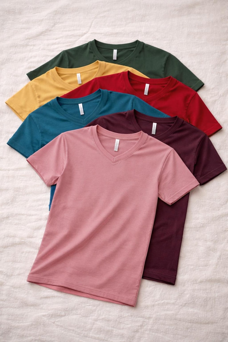 May include: A stack of six short-sleeved v-neck t-shirts in various solid colors including dark green, yellow, red, teal, maroon, and dusty pink. Each shirt features a white neck label and is made of a smooth, plain fabric with short sleeves.
