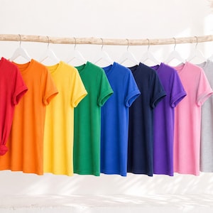 K&ouml;nnte beinhalten: Eine Kollektion von zehn kurz&auml;rmeligen T-Shirts mit Rundhalsausschnitt, die auf wei&szlig;en Kleiderb&uuml;geln in einer Reihe h&auml;ngen. Die T-Shirts sind in einem Farbspektrum aus Rot, Orange, Gelb, Gr&uuml;n, Blau, Dunkelblau, Lila, Pink, Grau und Wei&szlig; angeordnet.