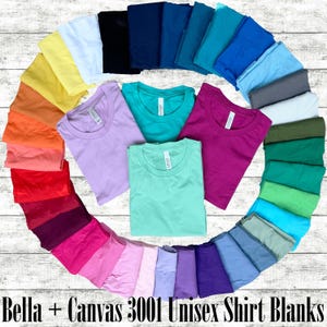 May include: A variety of Bella + Canvas 3001 unisex shirt blanks in different colors, arranged in a circle. The shirts are folded and displayed on a white wooden surface.