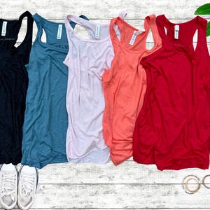 May include: A collection of six tank tops in various colors: black, teal, light pink, coral, and red. White sneakers and gold-toned glasses are also visible. The tank tops are arranged on a white wooden surface.