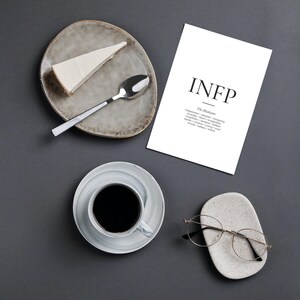 May include: Overhead shot of a serene arrangement on a dark gray surface. A slice of cheesecake sits on a speckled plate with a spoon. A cup of coffee, a print with "INFP" text, and round-framed glasses on a stone holder complete the scene.
