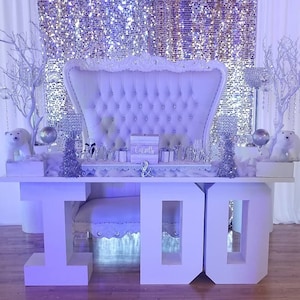 May include: A white, tufted, throne-like chair sits in front of a silver sparkly backdrop.  Two large white letters, "I" and "DO", are on a white table in front of the chair.  The table is decorated with white flowers and a white box with the word "Cards" on it.