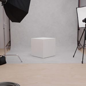 May include: A white cube sits on a gray surface in a photography studio. A black softbox and a camera on a tripod are positioned on either side of the cube. A black umbrella light and a coiled gray cable are also visible.