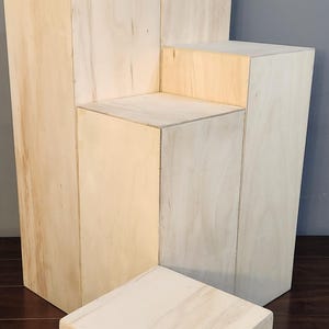 Puede incluir: Un conjunto de cuatro elevadores de exhibici&oacute;n de madera en varios tama&ntilde;os, hechos de madera de color claro con un acabado natural. Los elevadores est&aacute;n dispuestos en una formaci&oacute;n escalonada, creando una plataforma de exhibici&oacute;n en niveles.