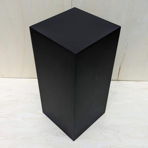 May include: A tall, black, geometric pedestal with a square top and a tapered design. The pedestal is made of a solid material and has a matte finish. The object is set against a light-colored wooden background.