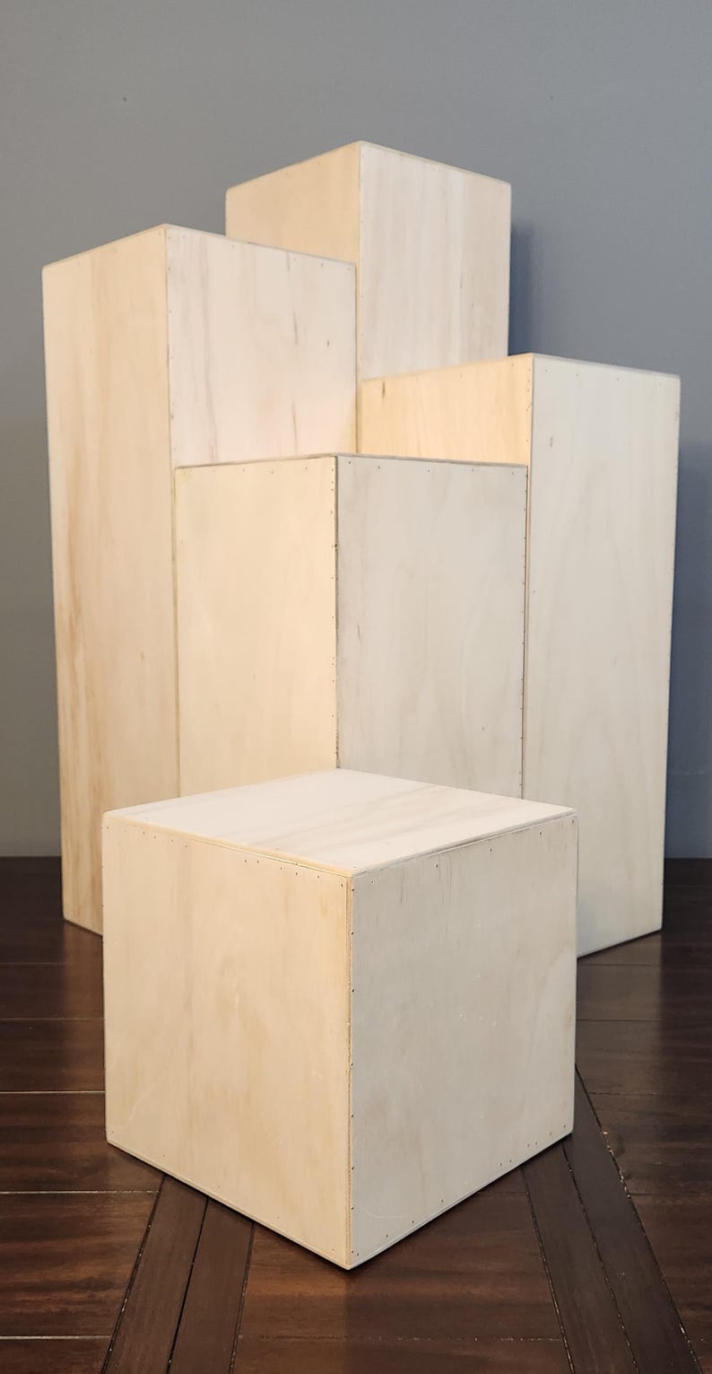 Puede incluir: Cinco elevadores de exhibici&oacute;n de madera de diferentes tama&ntilde;os, apilados en forma de pir&aacute;mide. Los elevadores est&aacute;n hechos de madera de color claro y tienen una superficie lisa y sin terminar.