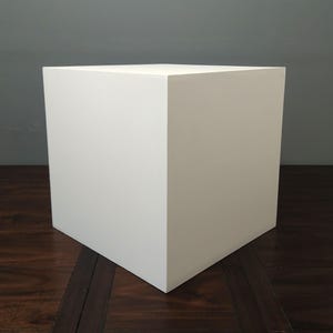 May include: A white, square cube with sharp corners. The cube is positioned on a dark wood surface. The object is a solid, geometric shape, and the lighting creates subtle shadows on its sides.