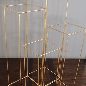 May include: A set of four gold metal square stands of varying sizes. The stands are made of thin metal and are designed to hold flowers or other decorative items.