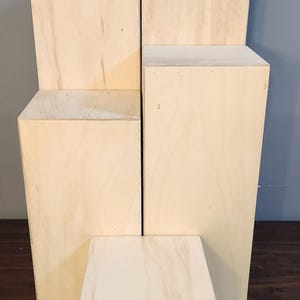 Puede incluir: Cuatro elevadores de exhibici&oacute;n de madera de diferentes tama&ntilde;os, apilados uno encima del otro. Los elevadores est&aacute;n hechos de madera de color claro y tienen una superficie lisa y sin terminar.