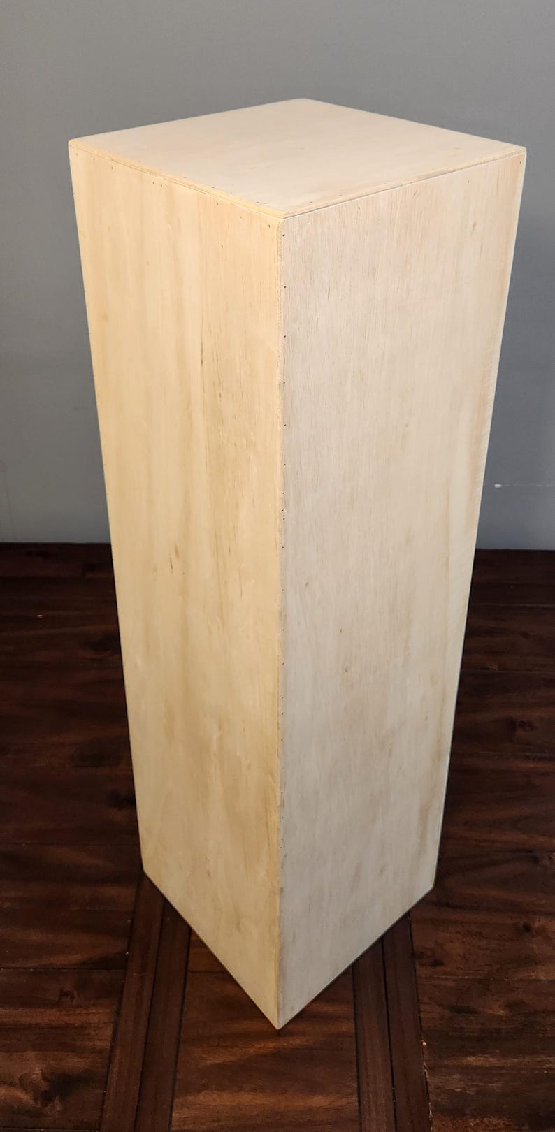 Puede incluir: Un pedestal rectangular de madera clara lisa con una superficie lisa. El pedestal est&aacute; sobre un suelo de madera oscura.