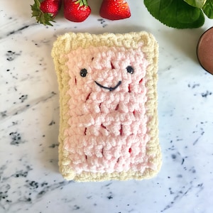 May include: A handmade, plush, smiling Pop-Tart in a light pink filling with a beige crust. The Pop-Tart has black button eyes and a black stitched smile. Red accents are visible within the pink filling. Strawberries are in the background.
