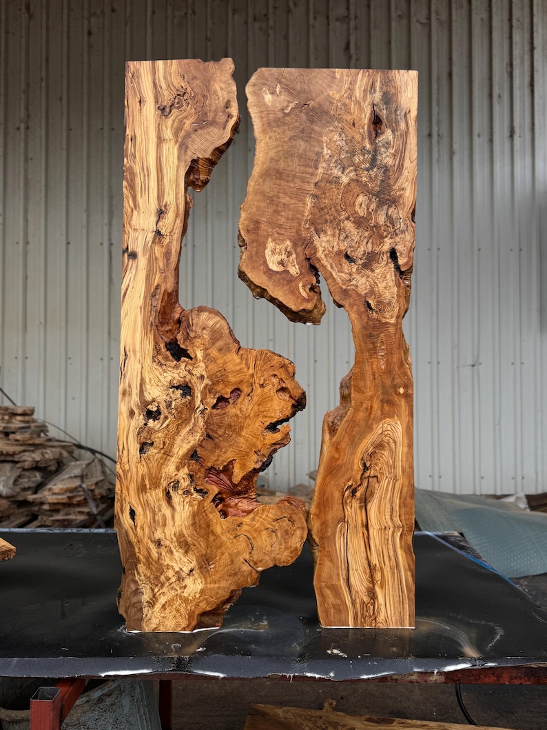 May include: Two vertical, unfinished wooden slabs with irregular edges and a natural, organic appearance. The wood has a warm, brown color with intricate grain patterns and knots, creating a rustic and unique aesthetic. The slabs are likely for a table or other furniture.
