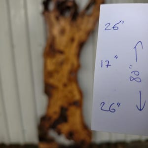 May include: A large, irregular live edge wood slab with a natural brown grain and dark knots. A white paper sign is attached, displaying the handwritten measurements of 26 inches, 17 inches, and 80 inches with directional arrows indicating the dimensions.