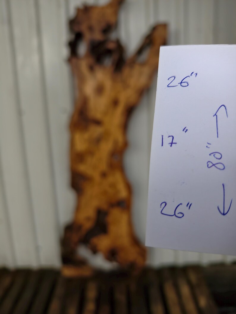 May include: A large, irregular live edge wood slab with a natural brown grain and dark knots. A white paper sign is attached, displaying the handwritten measurements of 26 inches, 17 inches, and 80 inches with directional arrows indicating the dimensions.