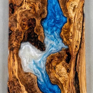 May include: A rectangular wooden art piece with a river-like design. The wood has a natural, warm brown color with intricate grain patterns. The river is made of blue and white resin, creating a flowing, water-like effect.