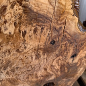 May include: A polished slab of light brown wood featuring a complex, swirling grain pattern with dark brown knots, organic textures, and natural fissures. The surface displays a high-gloss finish that highlights the intricate, marbled wood grain.