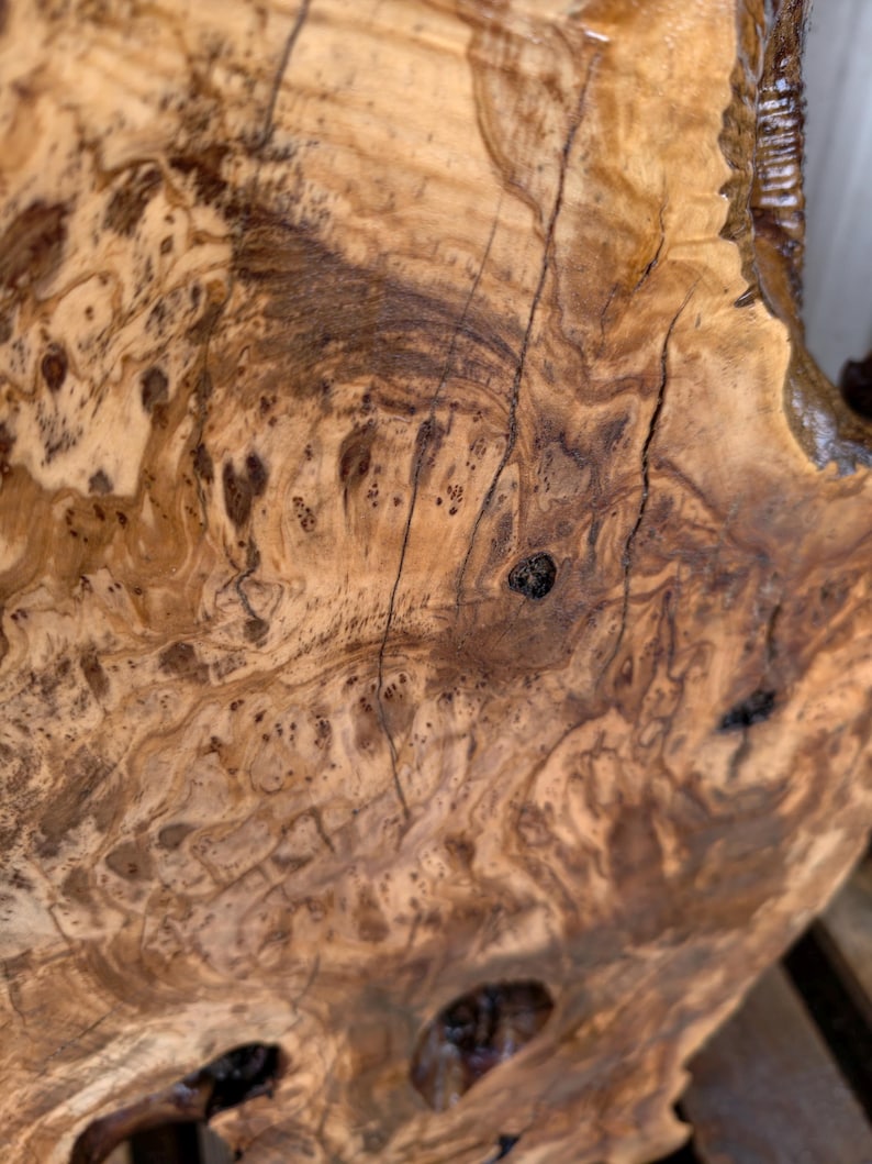 May include: A polished slab of light brown wood featuring a complex, swirling grain pattern with dark brown knots, organic textures, and natural fissures. The surface displays a high-gloss finish that highlights the intricate, marbled wood grain.